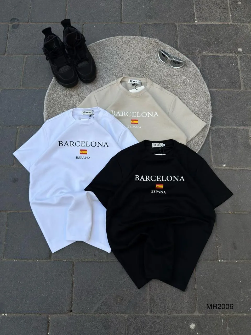 Barcelona Espana tees in white, sand and black laid flat on a round rug beside black sneakers