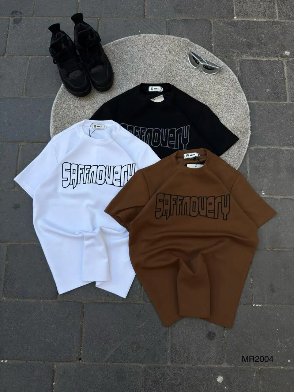 Outline logo tees in white, black and brown laid flat on cobblestones